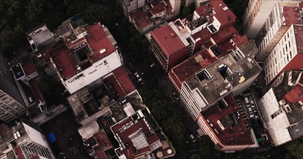 Bendita Suegra (2023) movie still: establishing — A dense urban district features multiple residential buildings with red tiled rooftops and…