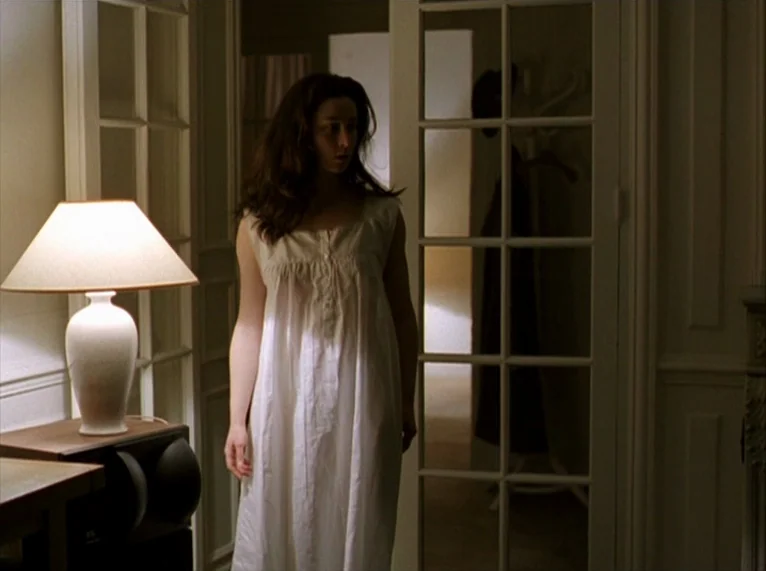 For Ever Mozart (1996) movie still: medium — A dimly lit hallway features a white lamp on a dark side table. A young woman in a white nightgown…