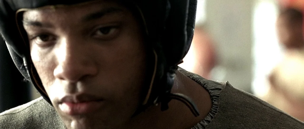Ali (2001) movie still: closeup — An athlete wearing headgear stands inside a training facility with blurry background elements. He looks intently…