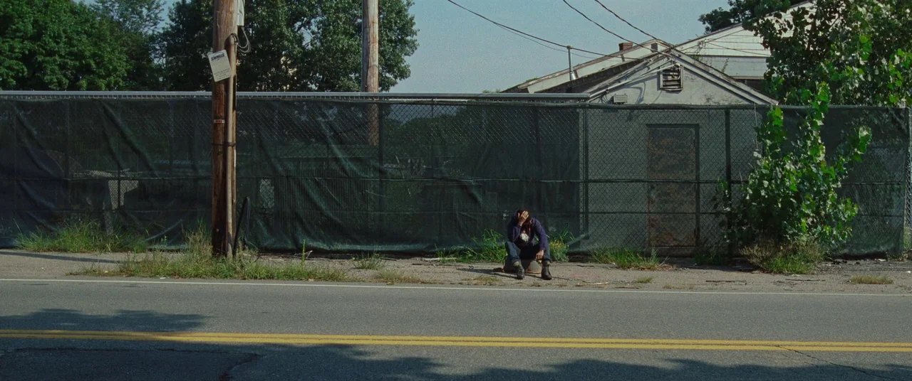 Sound of Metal (2019) movie still: wide — A roadside area features a chain-link fence covered in green mesh and a building. A lone person sits on a…
