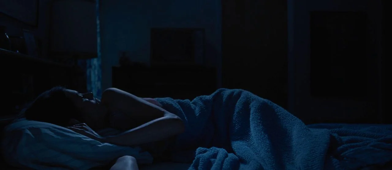 Seberg (2019) movie still: medium — A dark bedroom with minimal furniture silhouettes visible in the background. A person lies on their side in bed…