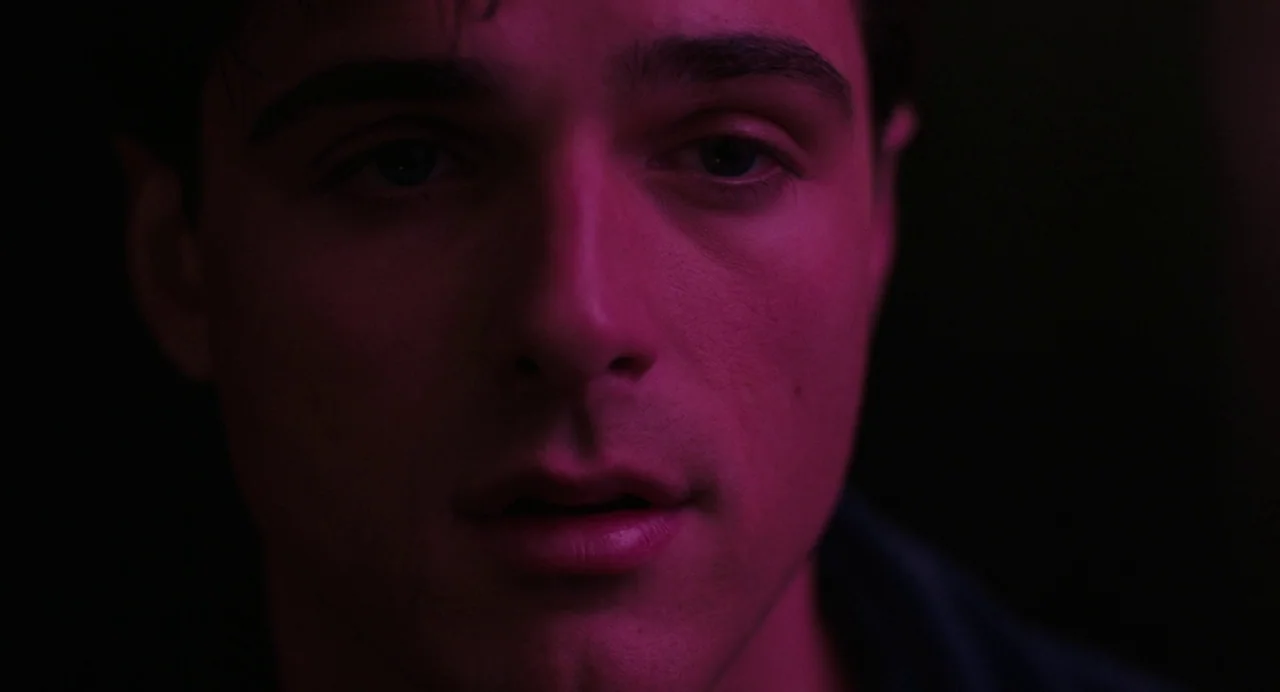 Priscilla (2023) movie still: closeup — A dark, indeterminate space is bathed in intense magenta light. A young man gazes directly into the camera…