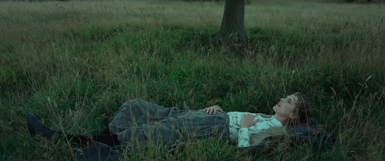 Sunset Song (2015) movie still: fullbody — A woman lies flat in a field of tall green grass under a large tree. She rests with her hands on her…