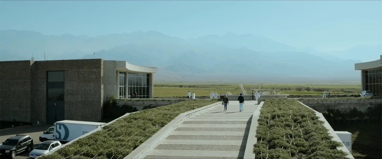 Checkmate (2015) movie still: wide — A modern architectural complex features a paved walkway leading toward an open mountain landscape. Two people…