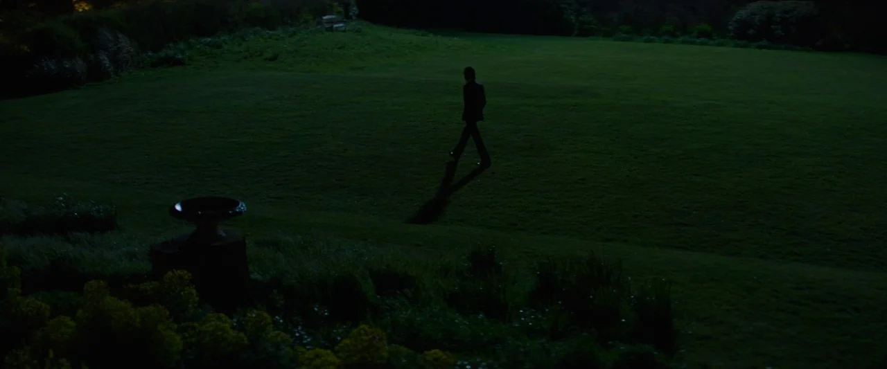 20,000 Days on Earth (2014) movie still: wide — A vast, dark lawn at night extends toward a distant, dimly lit boundary. A lone, silhouetted figure…