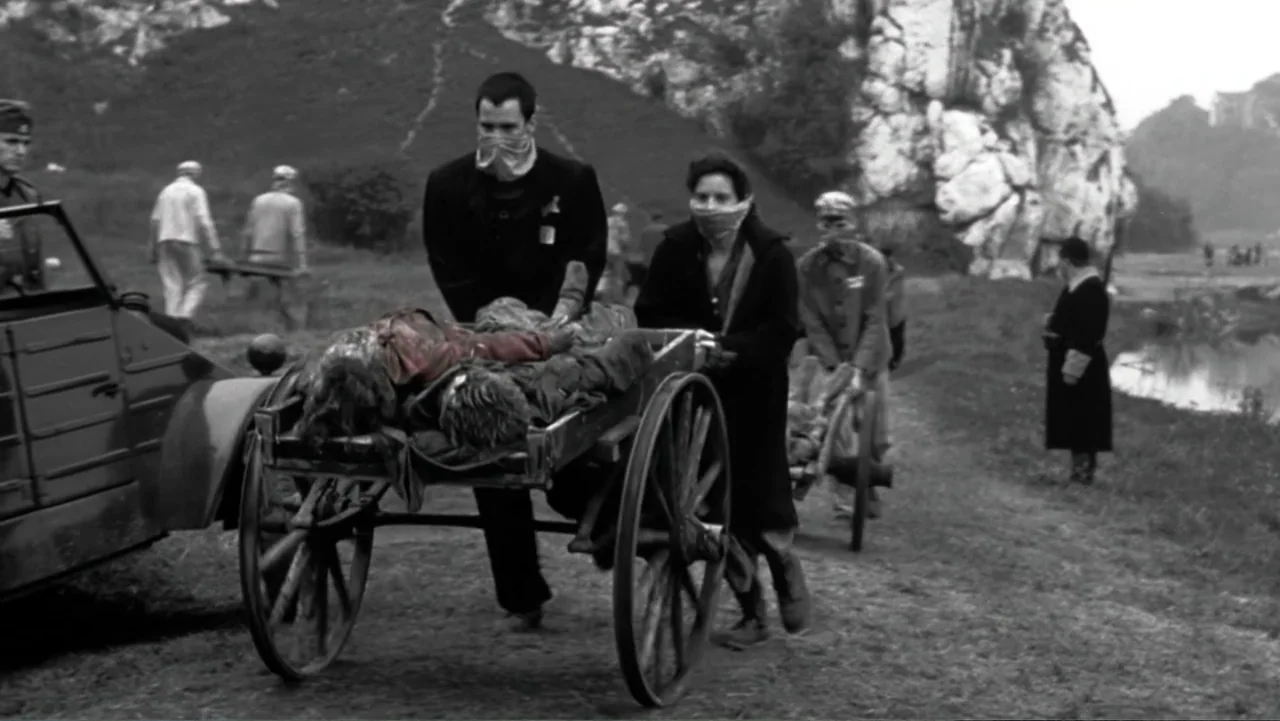 Schindler's List (1993) movie still: fullbody — A dirt path stretches through an open field with a large rocky outcrop. Two people push a wooden…