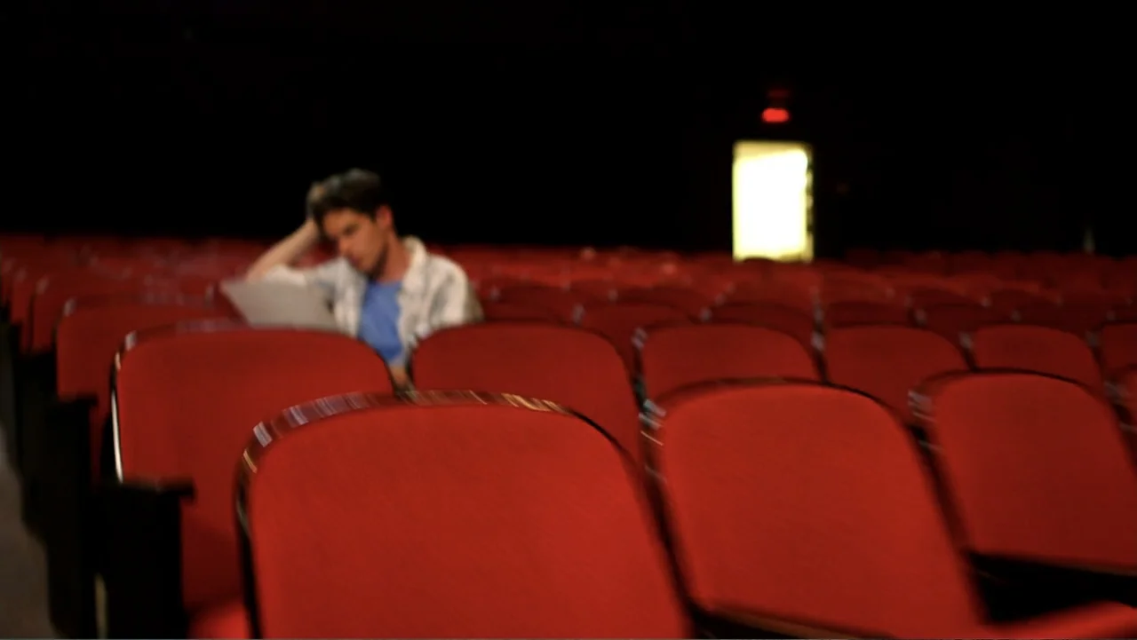 The Gallows (2015) movie still: medium — An empty movie theater features rows of red velvet seats in a dark room. A man sits alone in the mid-ground…
