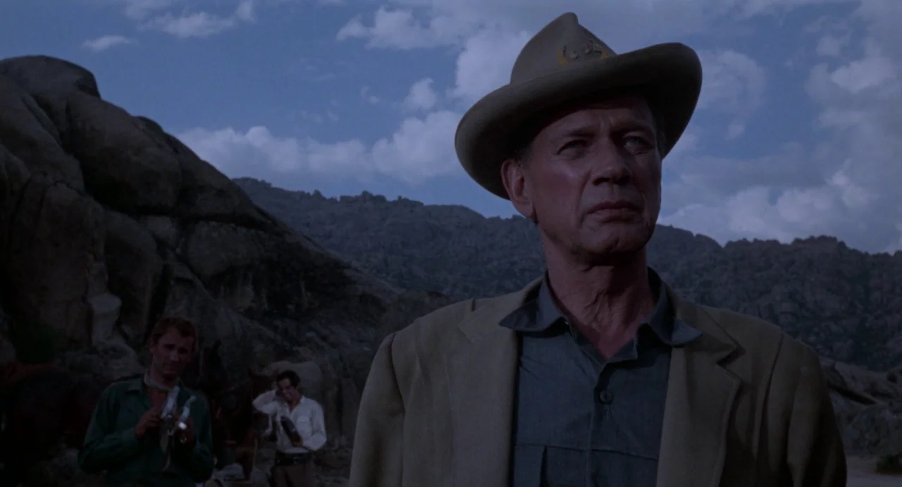 The Hellbenders (1967) movie still: medium — A group of men stands among large rock formations in a barren outdoor setting. The lead man in a tan…