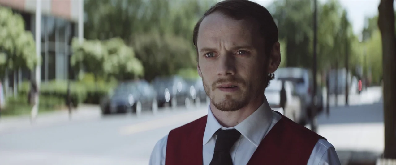 Thoroughbreds (2017) movie still: medium — A man stands on a blurred city sidewalk with trees and vehicles in background. The subject wears a formal…
