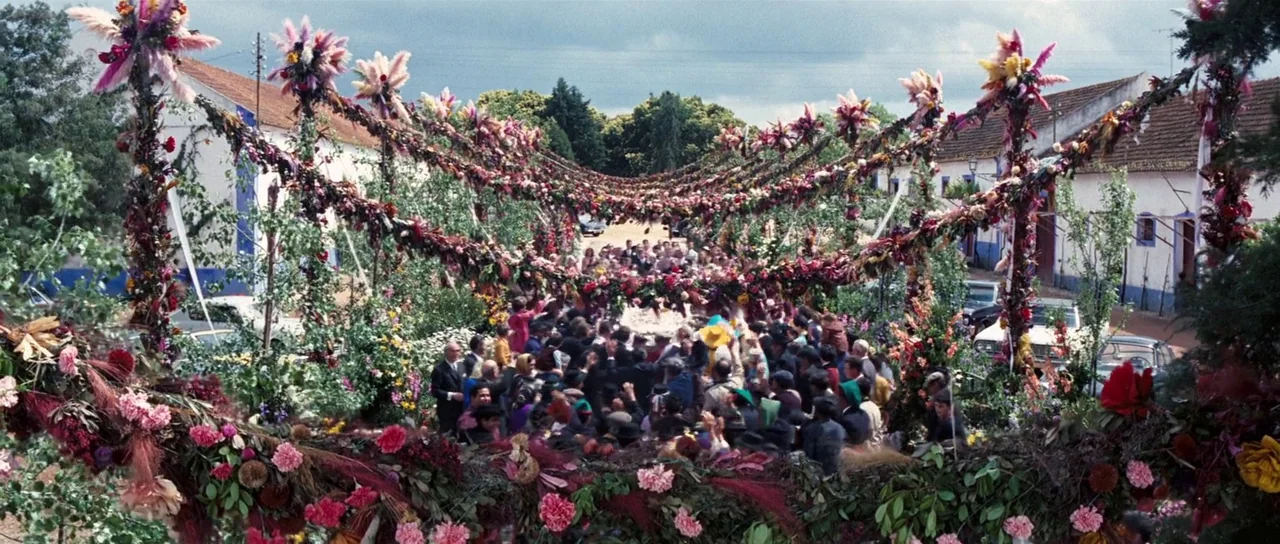 On Her Majesty’s Secret Service (1969) movie still: wide — A rural street is decorated with dense overhead garlands of colorful dried flowers. A…