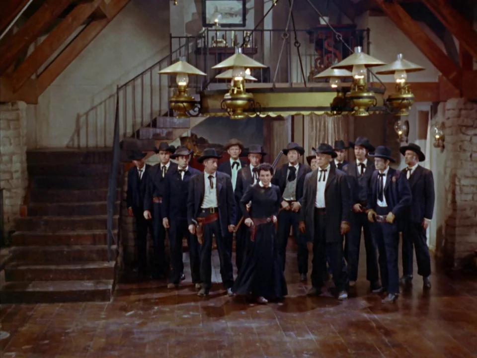 Johnny Guitar (1954) movie still: fullbody — A rustic timber-framed room features a central staircase and a large brass chandelier. A group of…