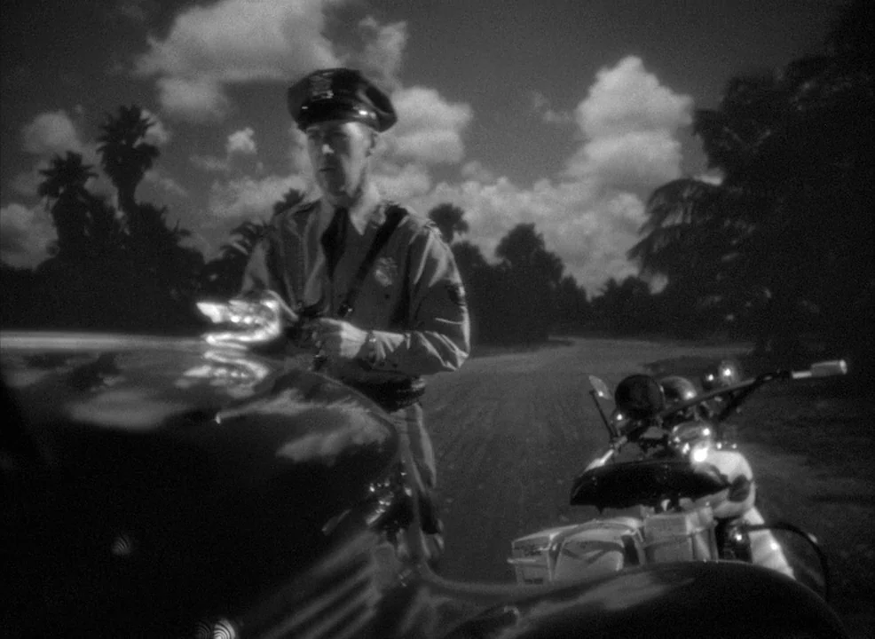 Notorious (1946) movie still: medium — A dark night scene shows a lone police officer standing on a dirt road. He examines the hood of a car parked…