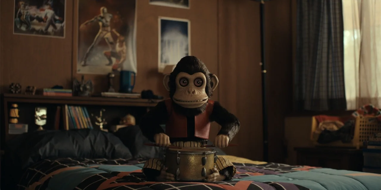 The Monkey (2025) movie still: medium — A dimly lit bedroom features wood-paneled walls decorated with faded vintage posters. A toy monkey in a red…