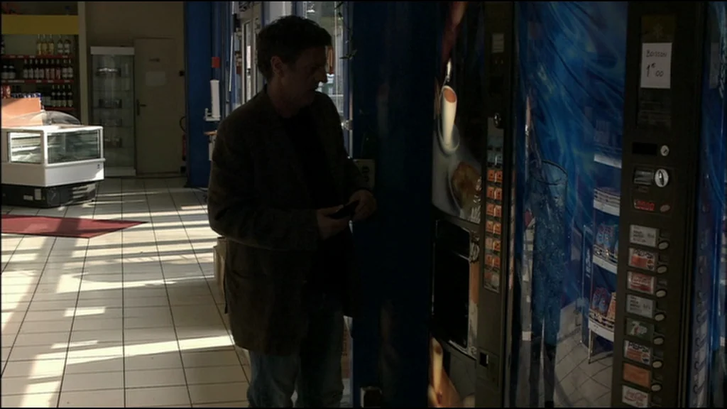 Caché (Hidden) (2005) movie still: medium — A tiled room contains a display freezer and a tall, blue-lit vending machine. A man stands in the aisle…