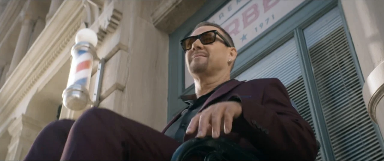 Man with No Past (2025) movie still: medium — A stone building facade features a spinning barber pole and a glass door storefront. A man in a…