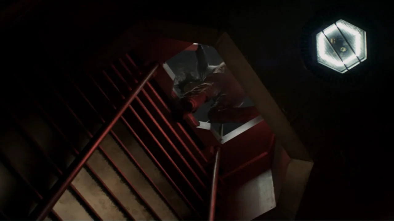 Total Recall (2012) movie still: wide — A dark industrial-style metal stairwell with a bright caged overhead light fixture. A lone figure in dark…
