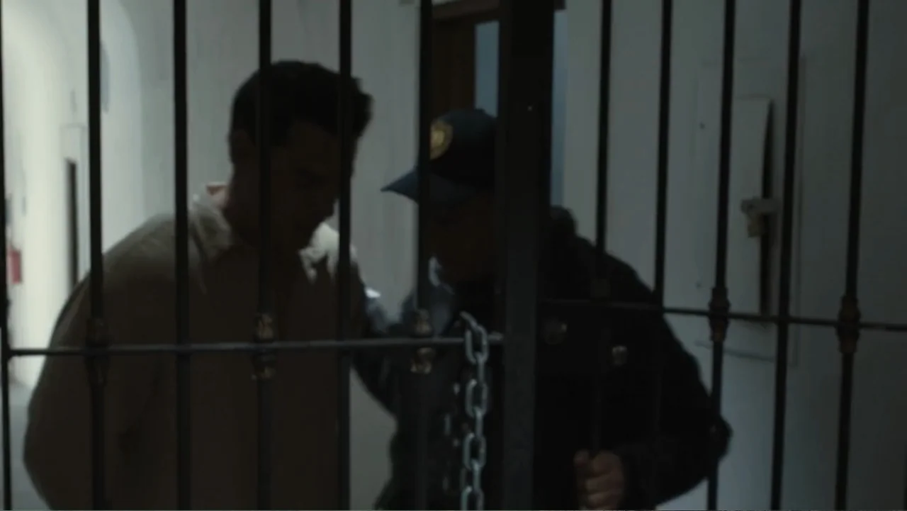 Por Amor al Money (2023) movie still: medium — A dimly lit interior space is obscured by vertical metal bars in the foreground. A man in a beige…