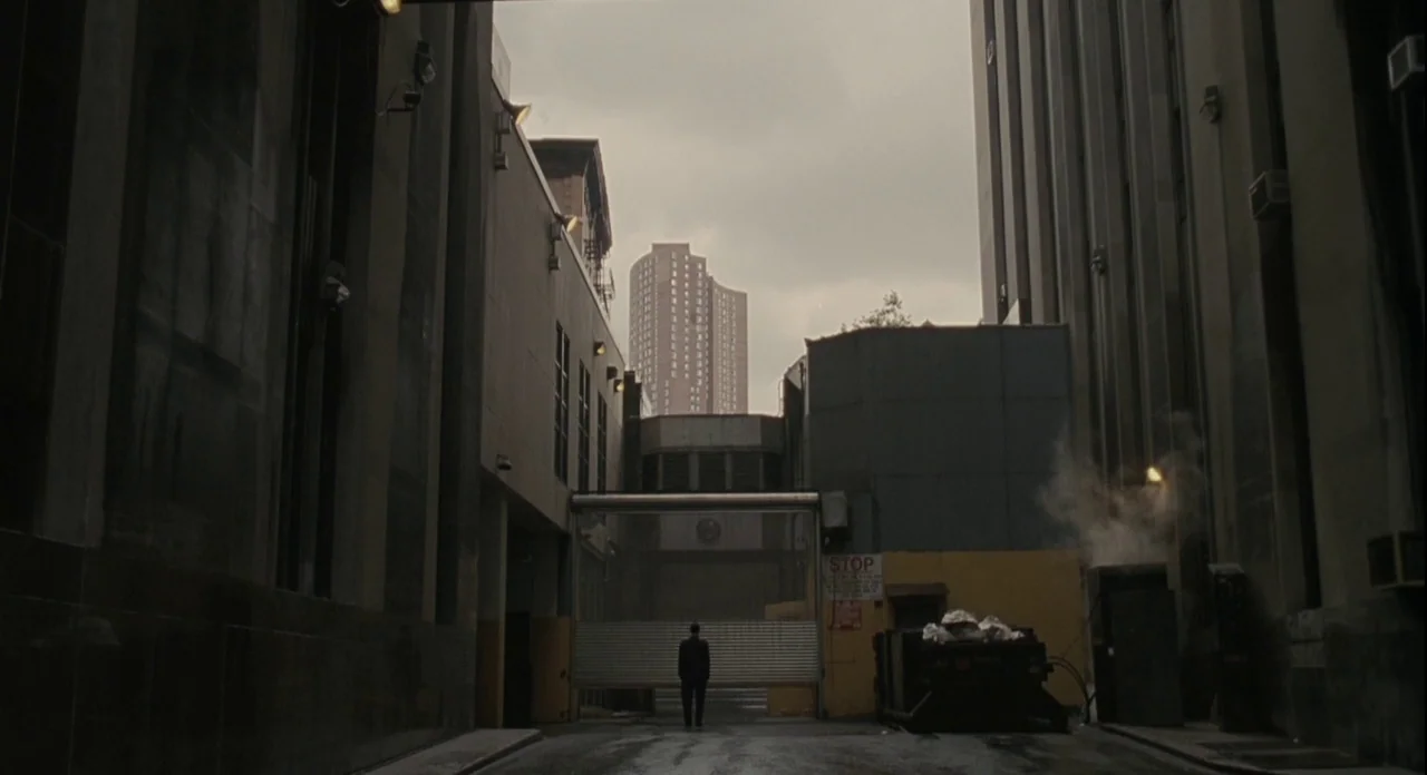 American Gangster (2007) movie still: wide — A narrow service corridor leads to a closed metal gate between tall concrete buildings. A lone man in a…