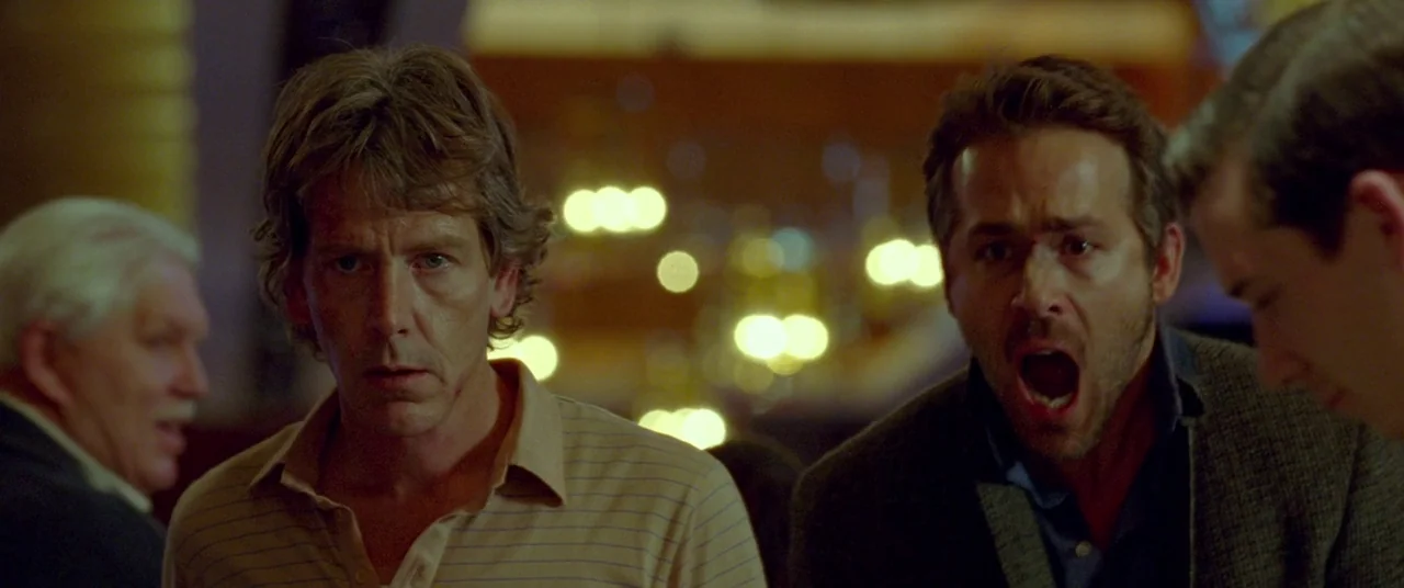 Mississippi Grind (2015) movie still: medium — A dimly lit indoor space features blurred golden bokeh lights in the deep background. Two men stand…
