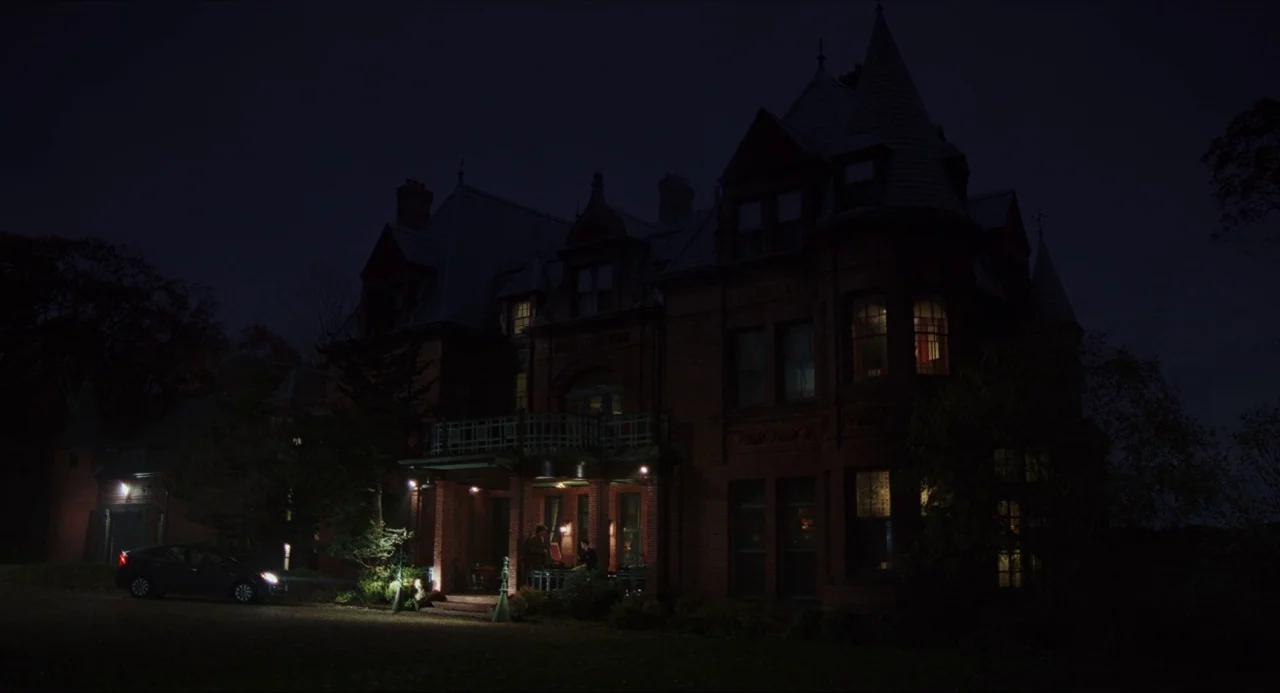 Knives Out (2019) movie still: wide — A dark brick Victorian mansion stands against a night sky with turreted rooflines. A lone dark car is parked…