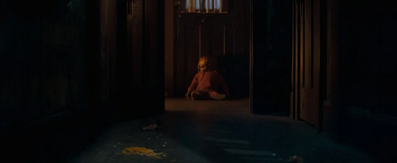 Trick ‘R Treat (2007) movie still: wide — A dark, shadowed hallway leads toward a closed wooden door with a curtained window. A small figure with a…