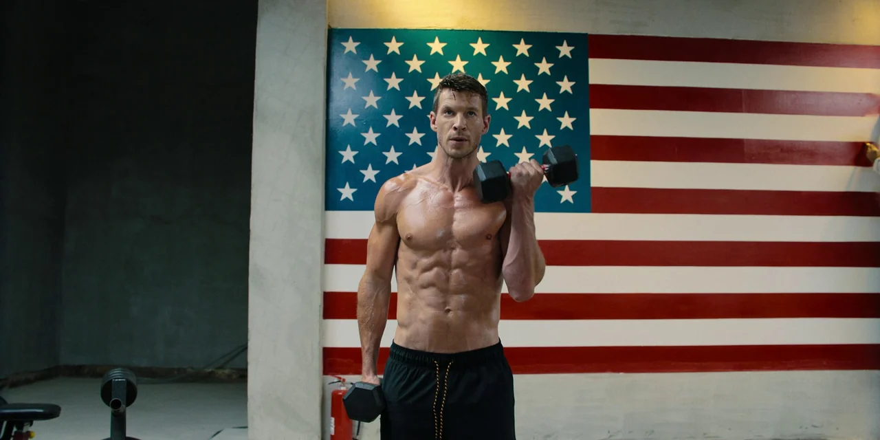 Sniper: G.R.I.T. (2023) movie still: medium — A man stands in front of a wall painted with an American flag. He performs a bicep curl holding a…