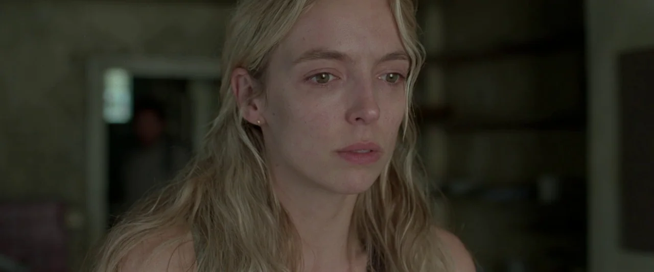 The End We Start From (2023) movie still: closeup — A dimly lit indoor space with muted earth tones and blurred background details. A woman with…