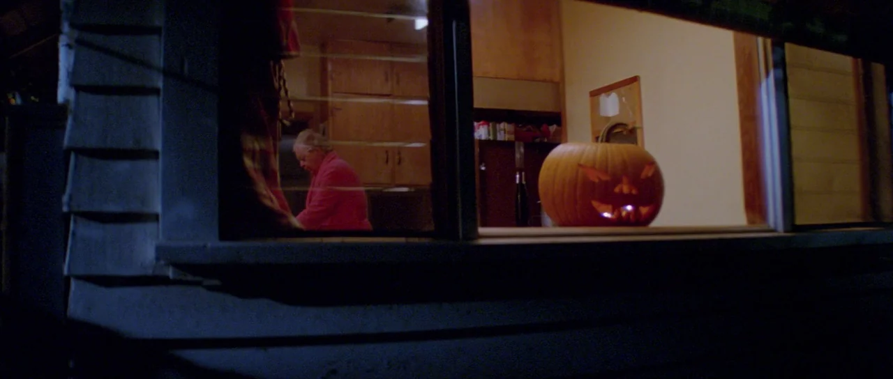 Halloween II (1981) movie still: wide — A dark house exterior shows an illuminated window looking into a dimly lit room. A carved jack-o'-lantern…