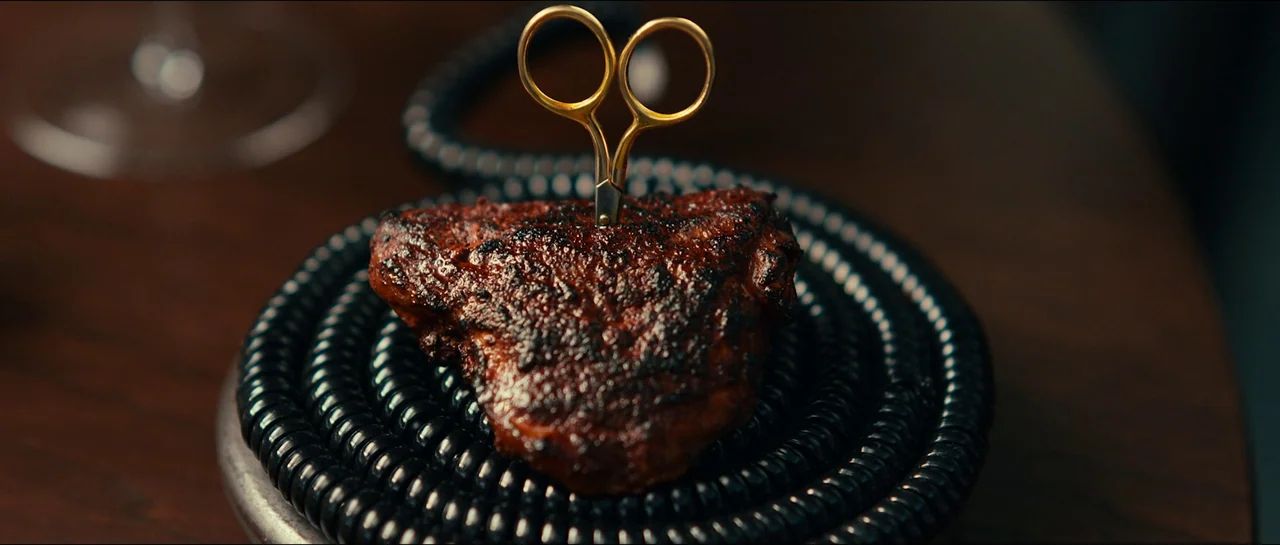 The Menu (2022) movie still: closeup — A piece of charred, cooked meat sits on a coiled black cable. Gold scissors are embedded upright in the…