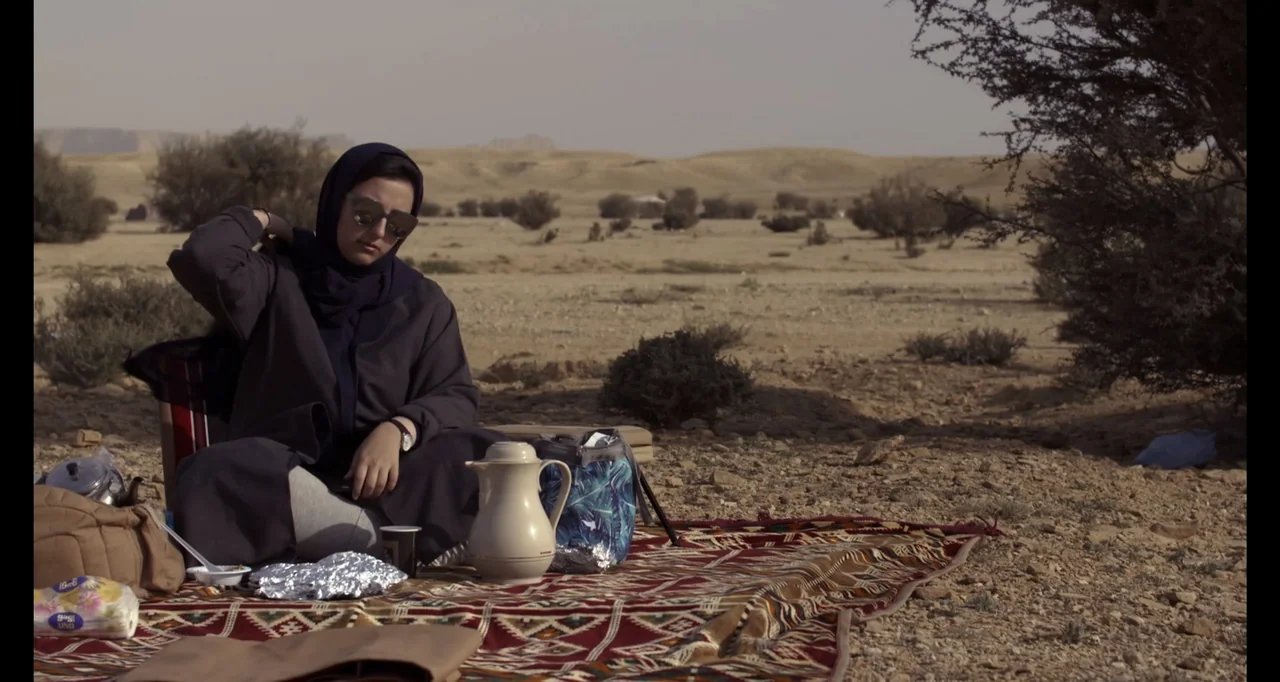 Saudi Women's Driving School (2019) movie still: medium — A dry desert landscape with sparse scrub bushes and a patterned rug laid out. A young…