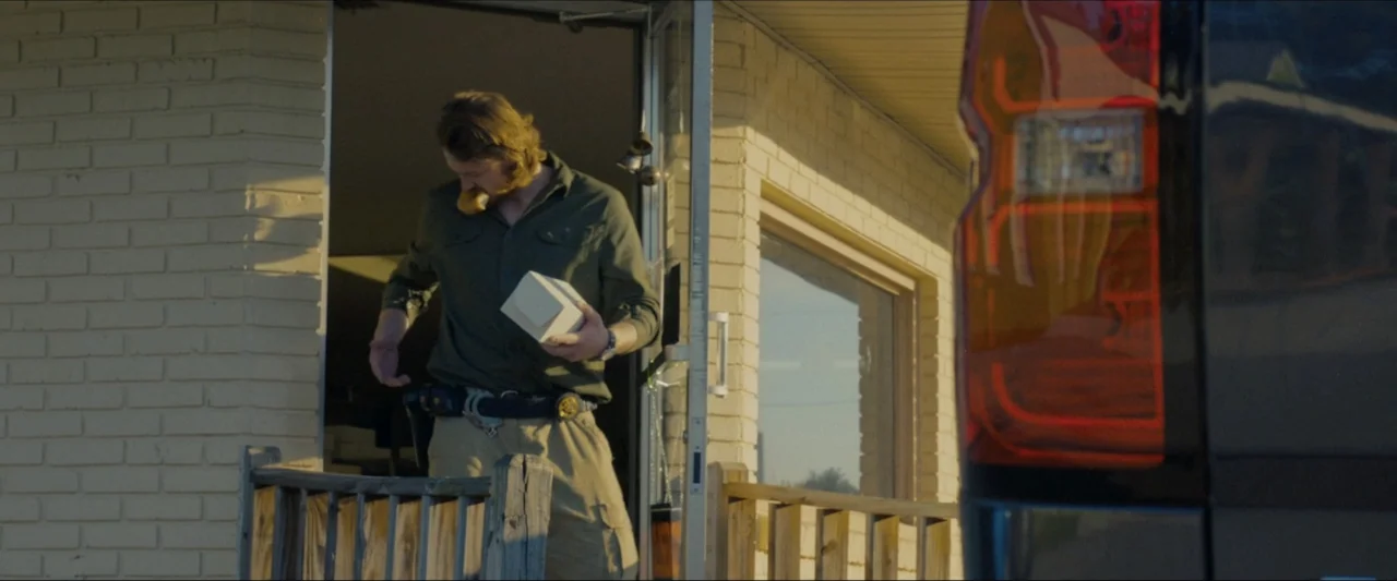 Ambush (2020) movie still: medium — A light-colored brick building exterior features an open doorway at golden hour. A man in a green shirt emerges…