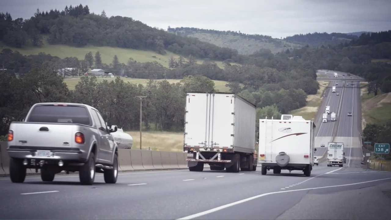 Beware of Angels (2016) movie still: wide — A multi-lane highway stretches through green hills under an overcast sky. Several trucks and passenger…