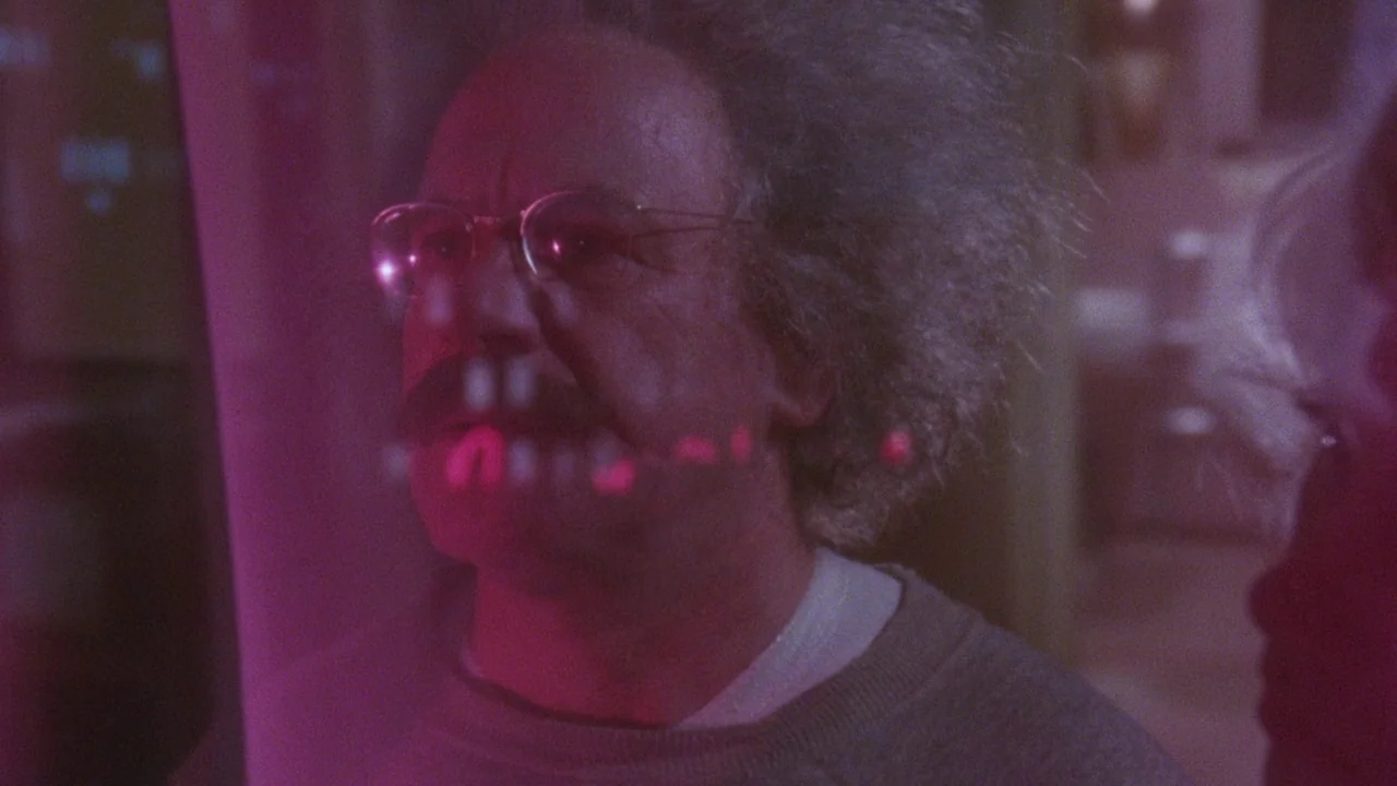 Insignificance (1985) movie still: closeup — An interior space with a magenta-tinted foreground reflecting off a glass surface. A man with wild hair…