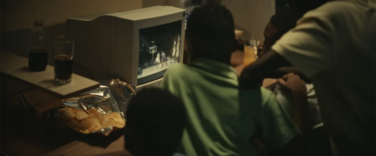 Giannis: The Marvelous Journey (2024) movie still: medium — A dimly lit room contains a desk with a vintage boxy computer monitor. Several children…