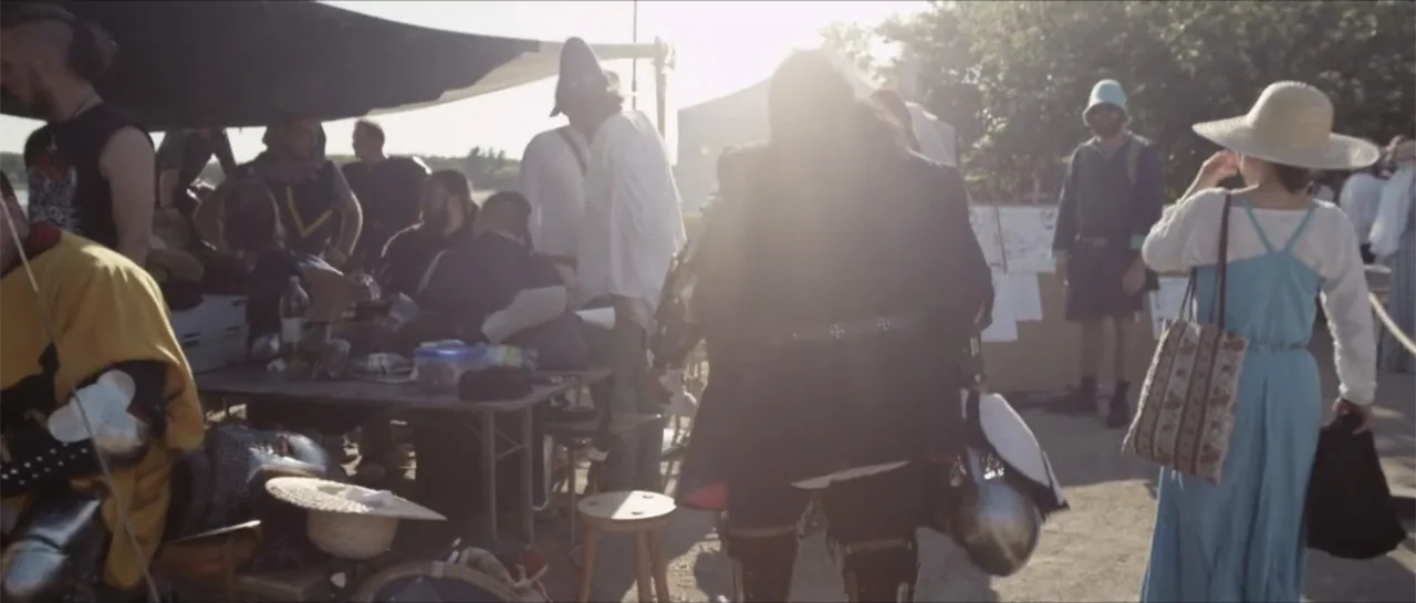 Bludgeon (2018) movie still: wide — A group of people wearing medieval-style clothing gather at an outdoor reenactment site. They sit and stand…