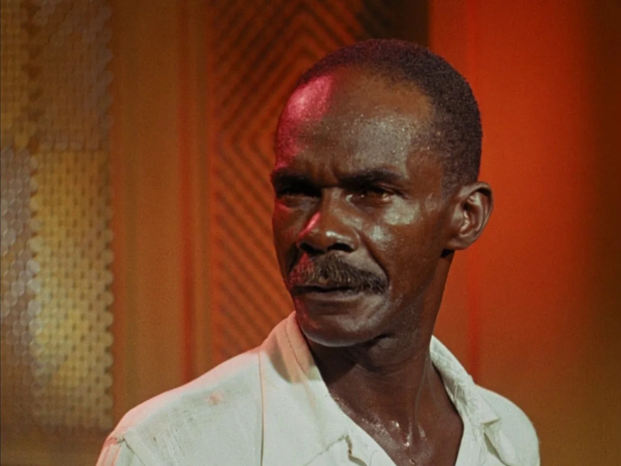 Black Orpheus (1959) movie still: closeup — A man stands indoors against a wall with patterned wood paneling and red lighting. The man, wearing a…