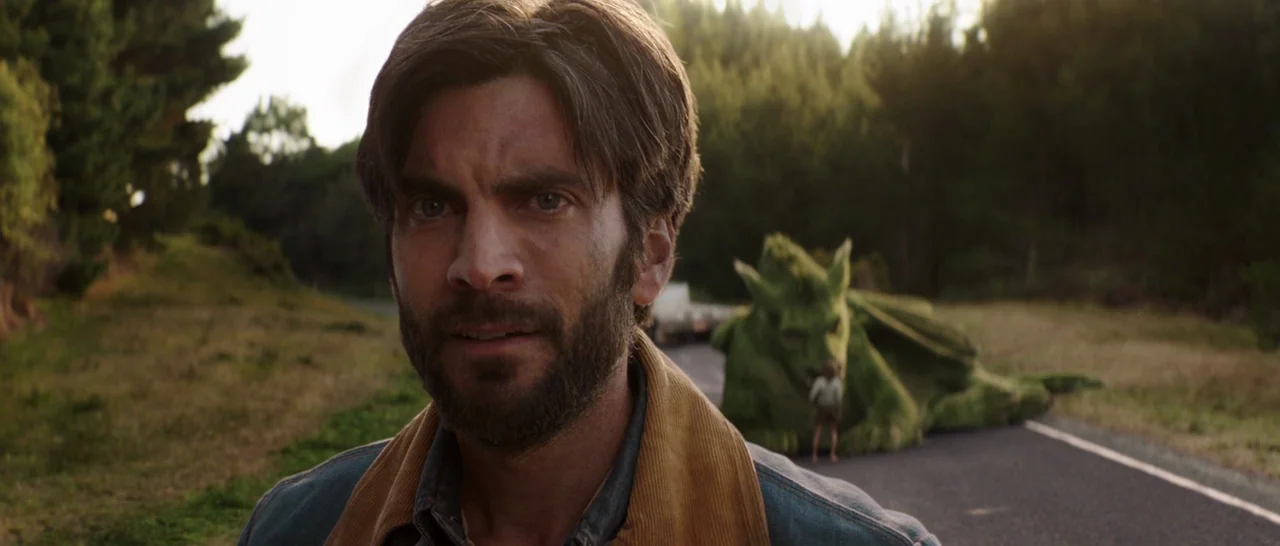 Pete’s Dragon (2016) movie still: medium — An asphalt road runs through a rural landscape surrounded by trees. A man looks at a large green…