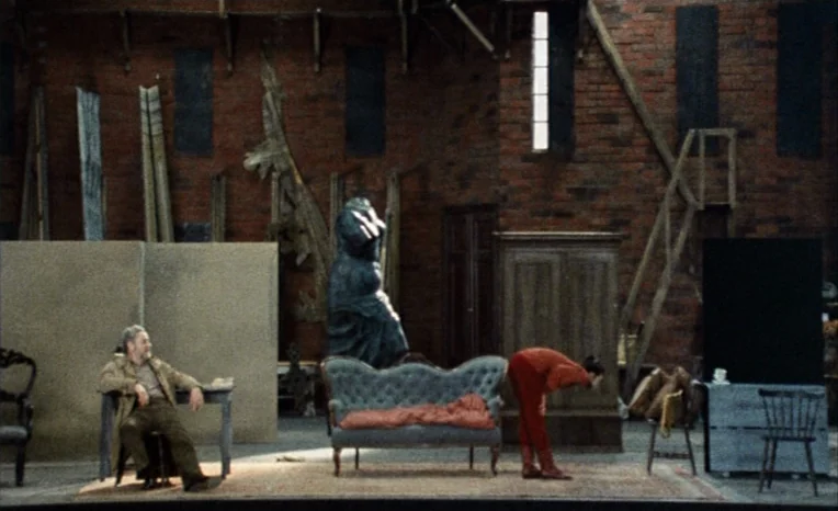 After The Rehearsal (1984) movie still: wide — A spacious room with high brick walls and various wooden panels and set pieces. An older man sits at…