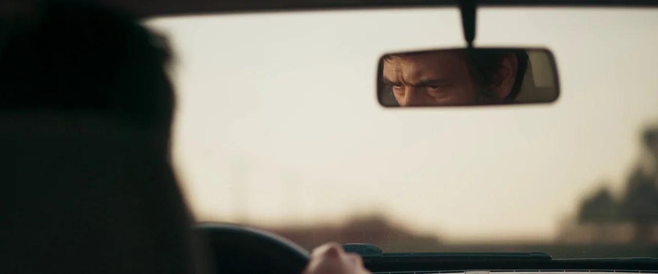 When Evil Lurks (2023) movie still: medium — The interior of a moving car shows a driver seen from behind. A man's face is reflected in the…