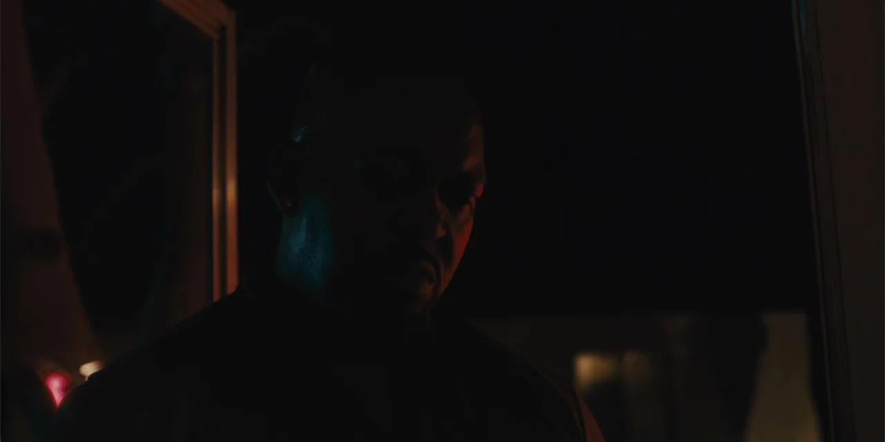 All Roads to Pearla (2019) movie still: closeup — A darkened interior doorway at night frames a man in low-key lighting. The man stands in the…