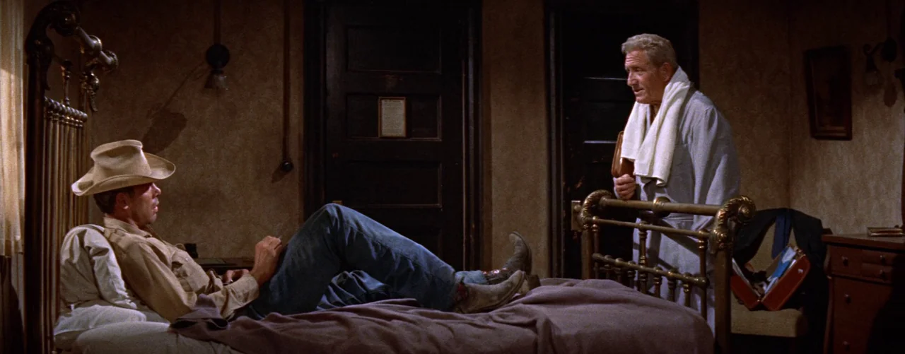 Bad Day At Black Rock (1955) movie still: wide — A dimly lit, dated motel room features dark wooden doors and muted wallpaper. A man in a cowboy hat…