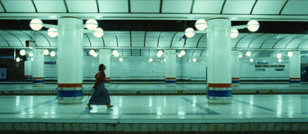 Brick Lane (2007) movie still: wide — A spacious, sterile underground transport terminal with tiled surfaces and bright overhead globe lights. A…