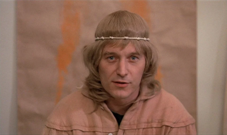 Lions Love (1969) movie still: closeup — A man stands in front of a tan wall with faint orange painted streaks. He looks towards the camera while…