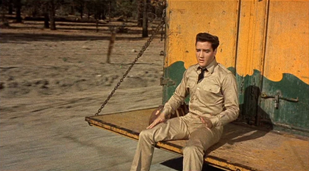Kid Galahad (1962) movie still: fullbody — A man sits on the tailgate of a yellow and green flatbed truck. The vehicle travels along a dirt road…
