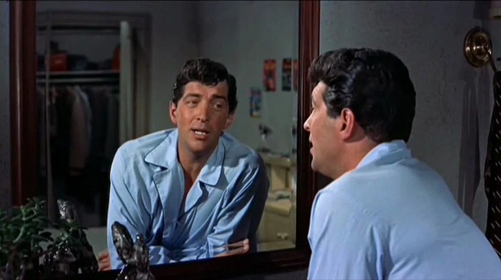 Artists and Models (1955) movie still: medium — A man stands in front of a framed mirror in a small dressing room. He watches his own reflection…