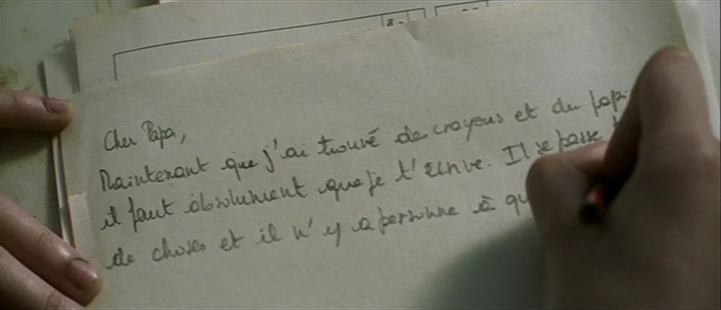 Time Of The Wolf (2003) movie still: closeup — A piece of aged stationery rests on a flat, pale surface. Someone holds a pencil while writing a…
