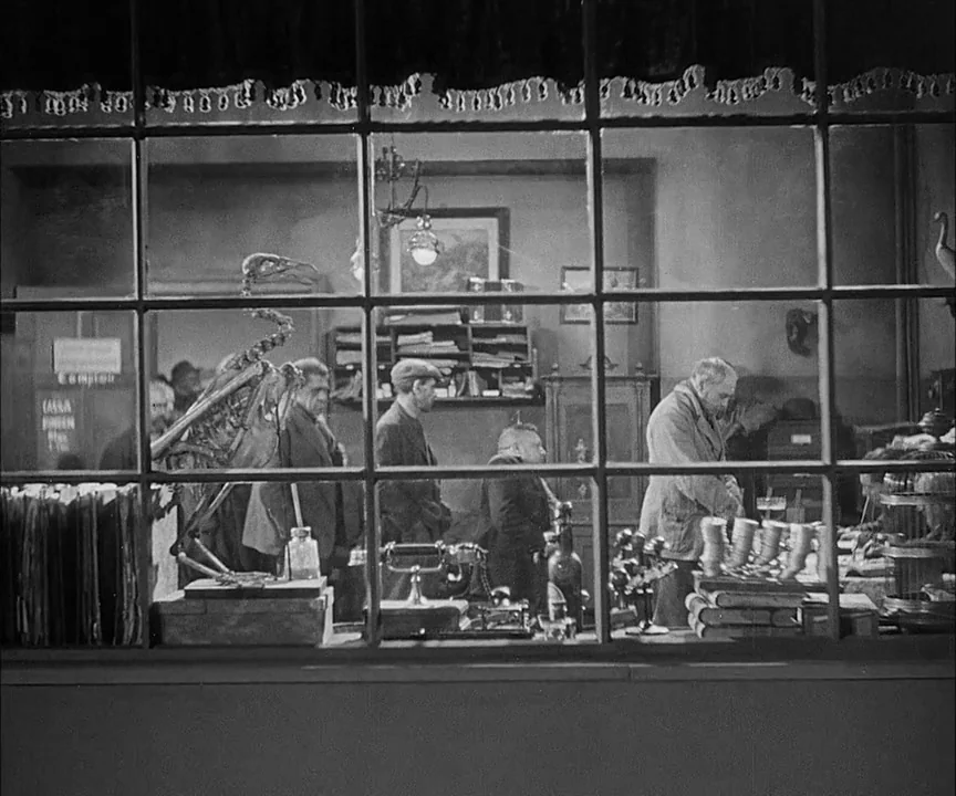 M (1931) movie still: wide — A cluttered shop interior viewed through a large multi-paned glass window at night. Several men stand at a counter…
