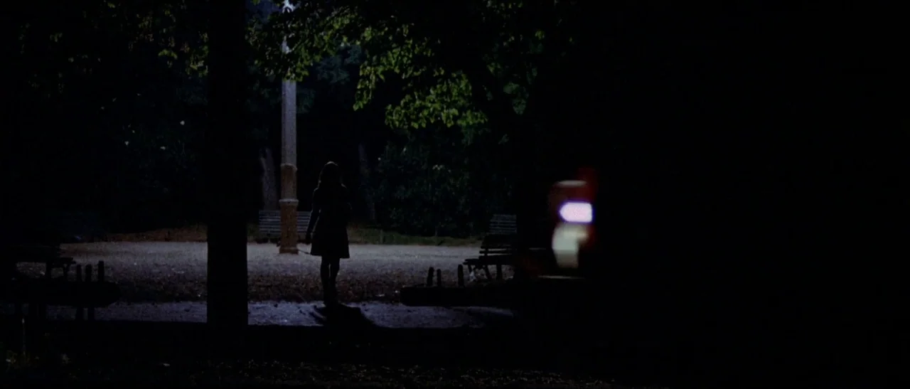 The Bird With The Crystal Plumage (1970) movie still: wide — A dark, empty city park at night is illuminated by a single streetlamp. A woman walks…
