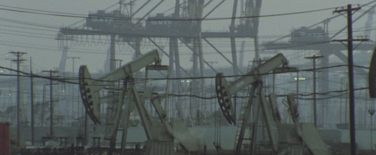 Syriana (2005) movie still: wide — Multiple oil pump jacks operate in a sprawling industrial field under a hazy sky. Large gantry cranes are visible…