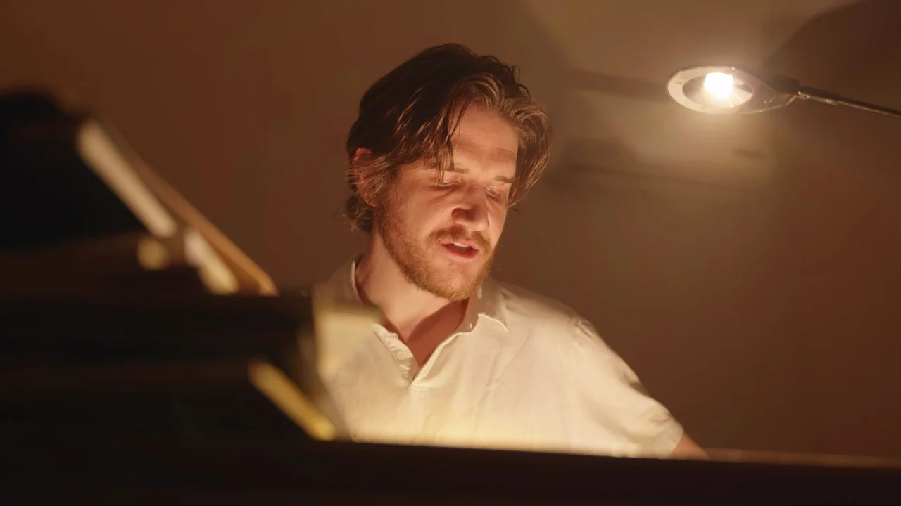 Bo Burnham: Inside (2021) movie still: medium — A man plays a piano in a dimly lit room with plain walls. He wears a plain white shirt while…